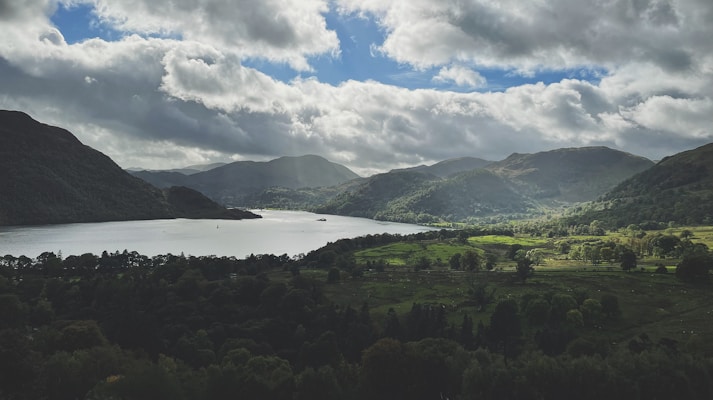 A serene landscape featuring a large body of water surrounded by rolling hills and mountains. The sky is filled with dramatic clouds, and there are patches of sunlight breaking through, illuminating parts of the green landscape. Trees and vegetation cover the area, enhancing the natural beauty.