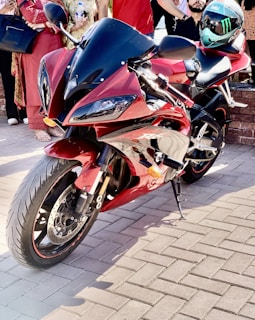 A sleek, red sports motorcycle parked on a paved surface. Several people stand nearby, some holding an object, possibly a bottle. A helmet is placed on the motorcycle's seat, featuring a distinctive design. The scene is well-lit, suggesting it is taken during the day.