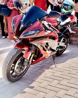 A sleek, red sports motorcycle parked on a paved surface. Several people stand nearby, some holding an object, possibly a bottle. A helmet is placed on the motorcycle's seat, featuring a distinctive design. The scene is well-lit, suggesting it is taken during the day.