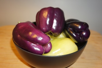 A black bowl contains several peppers, both purple and light yellow in color, arranged on a wooden surface. The peppers have a glossy texture and appear fresh.