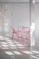 A minimalist interior space features a white brick wall, a white floor, and a set of red metal chairs surrounding a small white table. Sunlight streams through a window, casting shadows across the floor.