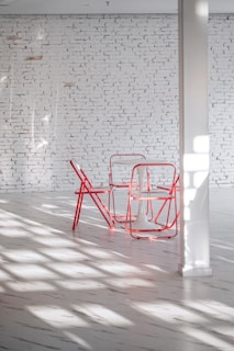 A minimalist interior space features a white brick wall, a white floor, and a set of red metal chairs surrounding a small white table. Sunlight streams through a window, casting shadows across the floor.
