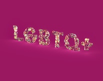 Glossy, multicolored letters spelling 'LGBTQ+' stand out against a solid magenta background. The letters have a reflective, shiny texture, creating a kaleidoscope effect with various shades.