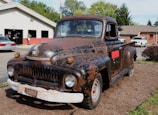 A rusty, vintage truck is parked outdoors on a bed of mulch in front of a service garage. The truck shows significant signs of wear, with faded and chipped paint revealing rusted metal beneath. In the background, there are several other vehicles and a building that appears to be an automotive repair shop.