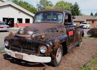 A rusty, vintage truck is parked outdoors on a bed of mulch in front of a service garage. The truck shows significant signs of wear, with faded and chipped paint revealing rusted metal beneath. In the background, there are several other vehicles and a building that appears to be an automotive repair shop.
