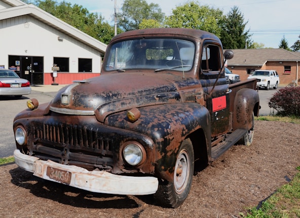 A rusty, vintage truck is parked outdoors on a bed of mulch in front of a service garage. The truck shows significant signs of wear, with faded and chipped paint revealing rusted metal beneath. In the background, there are several other vehicles and a building that appears to be an automotive repair shop.