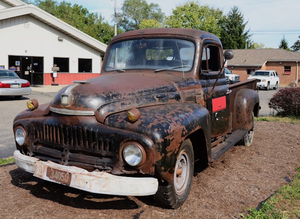A rusty, vintage truck is parked outdoors on a bed of mulch in front of a service garage. The truck shows significant signs of wear, with faded and chipped paint revealing rusted metal beneath. In the background, there are several other vehicles and a building that appears to be an automotive repair shop.