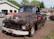 A rusty, vintage truck is parked outdoors on a bed of mulch in front of a service garage. The truck shows significant signs of wear, with faded and chipped paint revealing rusted metal beneath. In the background, there are several other vehicles and a building that appears to be an automotive repair shop.