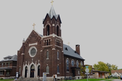 A large, historic church with a prominent brick façade and several pointed arches. It features multiple crosses on the roof and a tall steeple with a golden cross at its peak. Adjacent to the church are residential buildings and a street sign indicating a one-way street. Mature trees with autumn foliage are visible in the background.