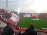 A large stadium filled with spectators features a vibrant soccer match atmosphere. Massive banners and flags are displayed in the stands on the left, while smoke in red and white colors creates a dramatic effect in the background. Players are lined up on one side of the field, and the stadium is packed with enthusiastic fans.