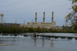 An industrial power plant with three tall smokestacks is situated behind a river with a small dam. The foreground features calm water and lush green trees surrounding the facility, with electrical transmission towers and power lines stretching into the distance. A cloudy sky casts a muted tone over the scene.