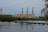 An industrial power plant with three tall smokestacks is situated behind a river with a small dam. The foreground features calm water and lush green trees surrounding the facility, with electrical transmission towers and power lines stretching into the distance. A cloudy sky casts a muted tone over the scene.