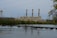 An industrial power plant with three tall smokestacks is situated behind a river with a small dam. The foreground features calm water and lush green trees surrounding the facility, with electrical transmission towers and power lines stretching into the distance. A cloudy sky casts a muted tone over the scene.