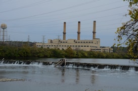 An industrial power plant with three tall smokestacks is situated behind a river with a small dam. The foreground features calm water and lush green trees surrounding the facility, with electrical transmission towers and power lines stretching into the distance. A cloudy sky casts a muted tone over the scene.