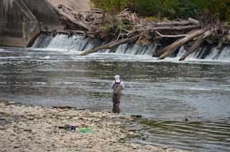 A person stands in a shallow river, facing a small waterfall created by logs blocking the flow of water. The individual is wearing fishing waders and a hat. The riverbank is covered with small pebbles and debris can be seen scattered along it. The background consists of lush green trees and more fallen logs near the waterfall.