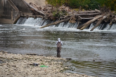 A person stands in a shallow river, facing a small waterfall created by logs blocking the flow of water. The individual is wearing fishing waders and a hat. The riverbank is covered with small pebbles and debris can be seen scattered along it. The background consists of lush green trees and more fallen logs near the waterfall.