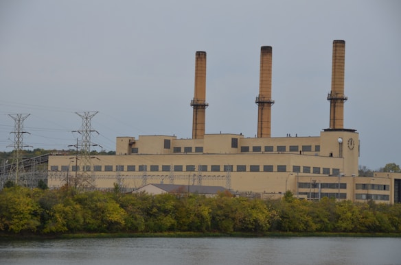 A large industrial building with multiple tall smokestacks is situated alongside a body of water. The building has a rectangular shape with numerous small windows and is surrounded by a line of trees. A series of electrical towers and power lines extend diagonally across the left part of the image, indicating an industrial setting.