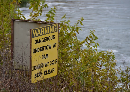 A weathered yellow warning sign amidst dense green foliage near a body of water. The sign warns about dangerous undertow conditions at a dam and advises people to stay clear.