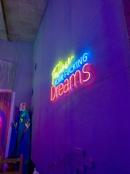 A vibrant neon sign with the text 'Follow Your F*cking Dreams' is mounted on a textured gray wall. The sign's colors include bright yellow, blue, and pink, casting a colorful glow. A decorated area with a surfboard, a beaded curtain, and a fire extinguisher sign is visible on the left.