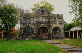 A large, two-story stone and stucco house with arched windows and a prominent central entrance surrounded by greenery. The structure combines rustic stonework with classical architectural features. The front yard is mostly grass, bordered by trees and an adjacent small structure.