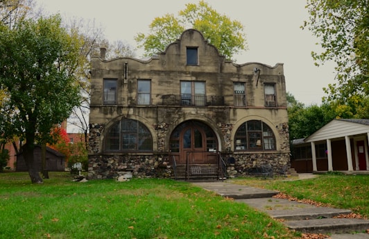 A large, two-story stone and stucco house with arched windows and a prominent central entrance surrounded by greenery. The structure combines rustic stonework with classical architectural features. The front yard is mostly grass, bordered by trees and an adjacent small structure.