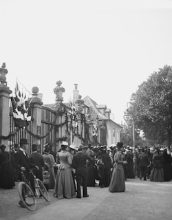 A large gathering of people in period attire, featuring a range of elaborate hats and dresses, is present near an ornate entrance adorned with flags and garlands. The scene is set outside a building with a distinct roof architecture and includes a few men with bicycles. Tall trees flank the area, contributing to the atmosphere of a formal event or celebration.