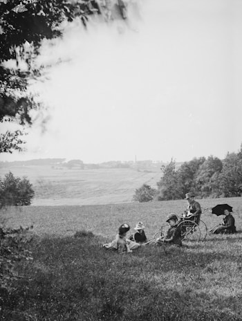 A group of children and a woman are relaxing on a grassy hill. One child is in a wheelchair while another holds an umbrella. The landscape in the background features rolling fields and scattered trees under a clear sky.
