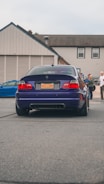 A dark blue BMW car is parked on a street. The car's license plate reads 'DAS BMR'. In the background, several people stand near a building with beige siding and a gray roof. Another vehicle, a blue car, is partially visible to the left.