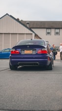 A dark blue BMW car is parked on a street. The car's license plate reads 'DAS BMR'. In the background, several people stand near a building with beige siding and a gray roof. Another vehicle, a blue car, is partially visible to the left.
