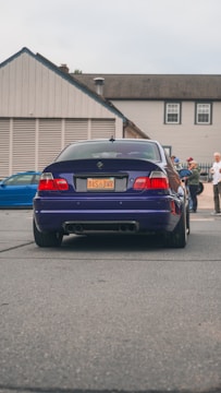 A dark blue BMW car is parked on a street. The car's license plate reads 'DAS BMR'. In the background, several people stand near a building with beige siding and a gray roof. Another vehicle, a blue car, is partially visible to the left.