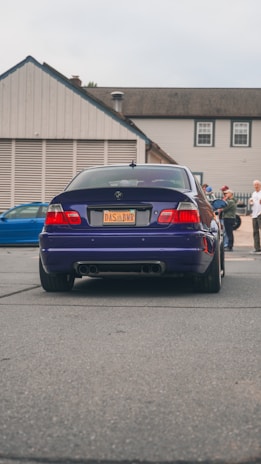 A dark blue BMW car is parked on a street. The car's license plate reads 'DAS BMR'. In the background, several people stand near a building with beige siding and a gray roof. Another vehicle, a blue car, is partially visible to the left.