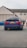 A dark blue BMW car is parked on a street. The car's license plate reads 'DAS BMR'. In the background, several people stand near a building with beige siding and a gray roof. Another vehicle, a blue car, is partially visible to the left.