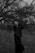 A person stands in a grassy field with their eyes closed and hands raised near their head, surrounded by bare trees under an overcast sky. The black and white image imparts a sense of solitude and introspection.