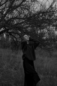 A person stands in a grassy field with their eyes closed and hands raised near their head, surrounded by bare trees under an overcast sky. The black and white image imparts a sense of solitude and introspection.