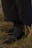 A pair of black leather loafers with silver buckles is worn with dark gray, textured socks. The footwear is partially covered by a long, dark garment, and the person is standing on dry, yellow and brown grass.