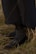 A pair of black leather loafers with silver buckles is worn with dark gray, textured socks. The footwear is partially covered by a long, dark garment, and the person is standing on dry, yellow and brown grass.