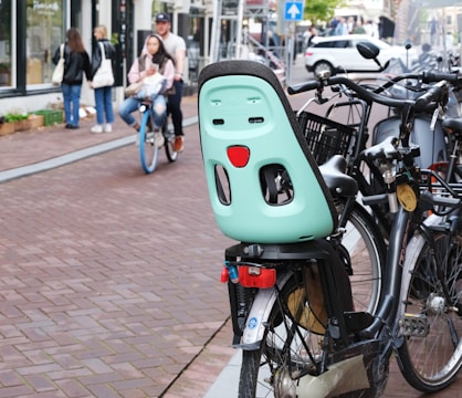 Several bicycles are parked on a brick paved street, with one prominently featuring a mint green child seat attached. In the background, a man and woman ride a bike together while a couple of pedestrians walk on the sidewalk. Cars are visible further down the street.