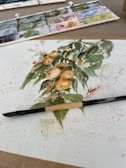 A watercolor painting depicting a bunch of oranges with green leaves on a white sheet of paper. The painting is surrounded by splatters of paint. A brush is placed across the lower part of the painting. A watercolor palette with various colors is seen in the background.