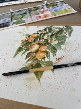 A watercolor painting depicting a bunch of oranges with green leaves on a white sheet of paper. The painting is surrounded by splatters of paint. A brush is placed across the lower part of the painting. A watercolor palette with various colors is seen in the background.