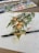A watercolor painting depicting a bunch of oranges with green leaves on a white sheet of paper. The painting is surrounded by splatters of paint. A brush is placed across the lower part of the painting. A watercolor palette with various colors is seen in the background.