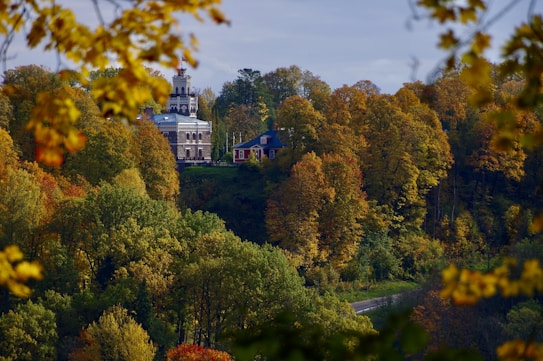 A picturesque autumn scene captures a grand building with a tower nestled among vibrant, colorful foliage. The leaves range from green to shades of yellow, orange, and red, signifying the fall season. A smaller house is located nearby, and a road cuts through the landscape at the bottom.