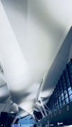 An architectural view of an airport terminal ceiling featuring modern design elements with white structural supports and large glass windows allowing natural light to enter. The ceiling has an intricate pattern and expansive space.