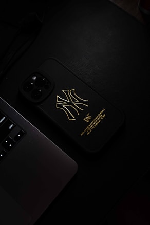 A dark, moody photograph featuring a smartphone with a black case that has a gold logo on it, placed next to the keyboard section of a laptop. The image has a minimalistic and sleek aesthetic, with a focus on the texture and contrast between the black case and the golden logo.