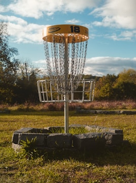 A disc golf basket standing in a grassy field, surrounded by a circular arrangement of stones. The basket is equipped with chains and a yellow top marked with the number 18. In the background, there are trees and a partly cloudy sky, indicating a pleasant outdoor setting.