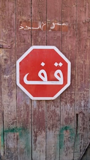 A red stop sign with white Arabic text is mounted on a weathered wooden wall. The wall shows signs of age with peeling paint and visible texture. Above the sign, there is some faded lettering on the wood.