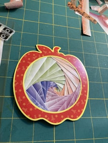 A paper craft project features a spiral pattern made from colorful folded paper, set within a red apple-shaped frame with yellow stars. The background consists of a grid-patterned green cutting mat scattered with various paper scraps.