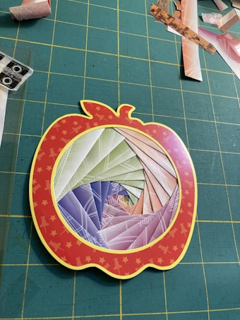 A paper craft project features a spiral pattern made from colorful folded paper, set within a red apple-shaped frame with yellow stars. The background consists of a grid-patterned green cutting mat scattered with various paper scraps.