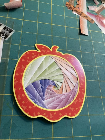 A paper craft project features a spiral pattern made from colorful folded paper, set within a red apple-shaped frame with yellow stars. The background consists of a grid-patterned green cutting mat scattered with various paper scraps.