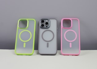 Three phone cases are displayed on a neutral background. The left case has a neon green border, the middle case contains a phone and has a gray border, while the right case features a pink border. Each case has a circular design in the center.