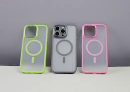 Three phone cases are displayed on a neutral background. The left case has a neon green border, the middle case contains a phone and has a gray border, while the right case features a pink border. Each case has a circular design in the center.